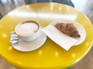 Freshly brewed coffee in white cup alongside flaky croissant on white plate, resting on vibrant yellow table, creating a cozy cafe atmosphere for breakfast enjoyment and relaxation