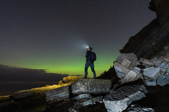 Adventurous person with a headlamp stands on a rocky cliff under a starry sky, watching the magical green northern lights. - Powered by Adobe