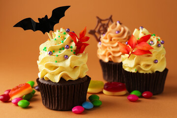 Delicious cupcakes with bat topper, branches with red leaves, sprinkles and candies on orange background, closeup. Halloween celebration