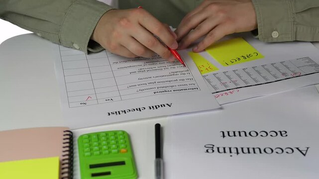 A man's hands in a formal shirt carefully review an audit checklist placed alongside a balance sheet with handwritten notes. Using a red marker, he authoritatively checks off items on the list
