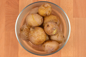 A glass bowl holds several whole boiled potatoes with their skins on, sitting on a light wooden surface