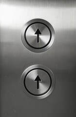 Close up view of two metal elevator buttons with up arrows. One button is slightly higher than the other. The brushed steel surface reflects light.
