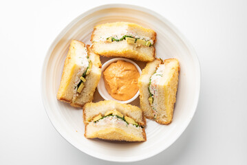 Assorted sandwich halves with cucumber, avocado, and creamy filling arranged around dipping sauce on white plate, top view, isolated on white background, suitable for food photography.