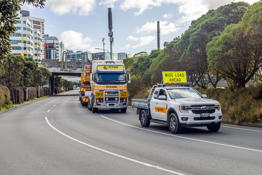 A white escort vehicle with a "Wide Load Ahead" sign leads an oversize Titan crane truck during the Wellington Crane Parade in New Zealand