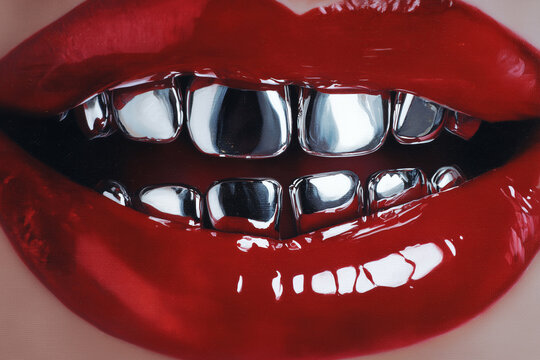 Close-up of glossy red lips revealing chrome dental grills, high gloss makeup and metallic teeth reflecting light in a bold fashion beauty portrait with dramatic shine.