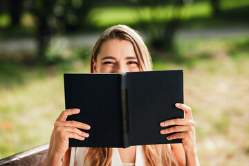 Obraz premium Young woman enjoys a sunny outdoor moment reading a black notebook while sitting on a park bench smiling and feeling relaxed in a bright spring day