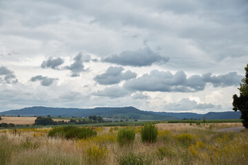 A panoramic view of a vast rural landscape with golden fields and distant forested hills under a dramatic, cloudy sky, evoking a sense of calm and nature's grandeur.