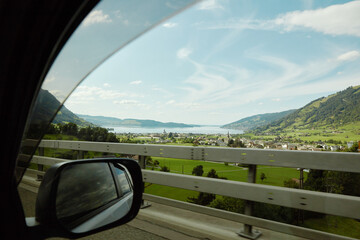 Scenic drive through a picturesque valley alongside a tranquil lake, framed by rolling green mountains and a charming village under a clear blue sky, viewed from a car window and side mirror.
