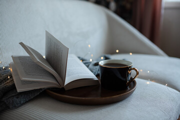 Cup of coffee with open paper book on wooden tray over Christmas lights and knitted sweater in couch closeup. Winter holiday season. Cozy home atmosphere.
