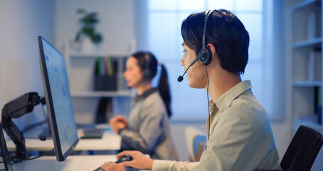 Asian male call center agent with headset working at night in a modern office, using computer for customer communication, IT support, business service and problem solving.