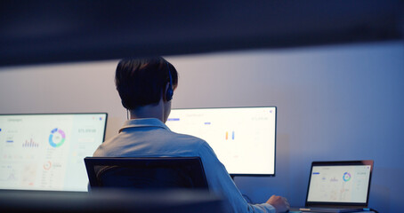 Rear view of Asian male call center service with headset working at night, using multiple computer screens for customer support, data analysis and communication in modern office.