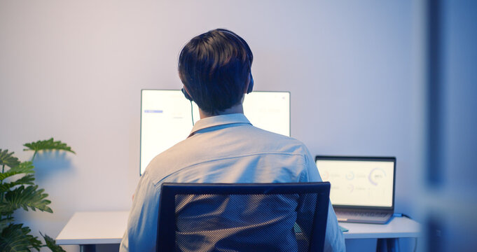 Back view asian male call center customer support with headset working late at night in modern office, using dual computer monitors for customer service, support and communication.