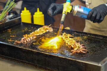Street food chef grilling squid skewers with a blowtorch.