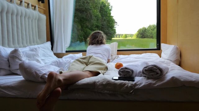 Young woman relaxing in a cozy bed while reading a book, with an orange magically floating beside her, overlooking a beautiful, green landscape through a large panoramic window
