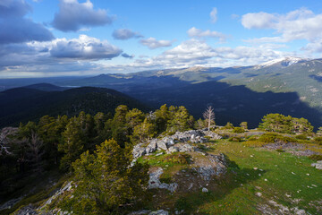 Rocky outcrop and pine forest at Cerro de la Camorca summit, Sierra de Guadarrama, Spain, with panoramic mountain views under vibrant blue sky and scattered clouds
