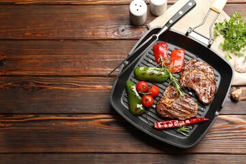 Pan with grilled beef steaks, rosemary, vegetables, shakers and carving fork on wooden table, flat lay. Space for text