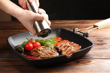 Woman adding pepper to grilled beef steaks, rosemary and vegetables in pan at wooden table against black background, closeup
