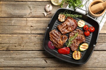 Pan with grilled beef steaks, rosemary and vegetables on wooden table, flat lay. Space for text