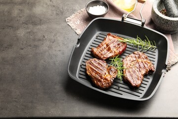 Pan with grilled beef steaks, rosemary and spices on grey textured table, closeup. Space for text