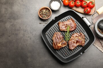 Pan with grilled beef steaks, rosemary, tomatoes and spices on grey textured table, flat lay. Space for text