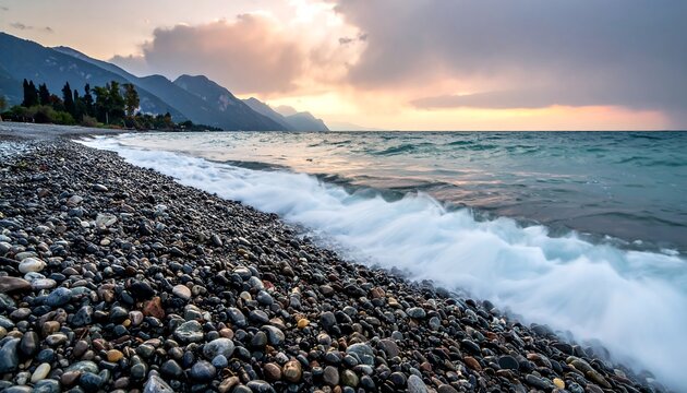 Coastal scene featuring a pebble beach with gentle waves rolling towards the shore, under a partly cloudy sky during the golden hour