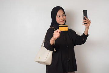 Cheerful Muslim woman wearing black hijab holding a credit card and smartphone while taking a selfie with confidence.