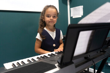 Portrait of a cute little girl kid playing digital keyboard electronic piano for up skill. Little artist musician learns the melody from the notes. Music studio or school.