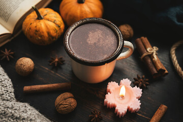 Cozy autumn scene with mug of hot chocolate, book, pumpkins, spices and candles on rustic dark wooden background, warm seasonal atmosphere