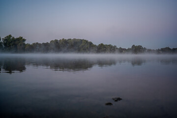 Ein ruhiger Morgen am Fluss mit Nebel und spiegelnder Wasseroberfläche in der Dämmerung