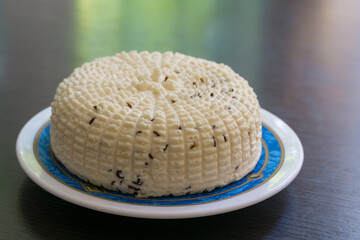 Round white homemade cheese with textured, ridged surface and   cumin spice, presented whole on decorative plate with blue and gold rim
