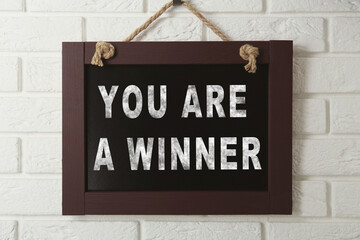 Fototapeta premium Chalkboard with phrase You Are A Winner hanging on white brick wall