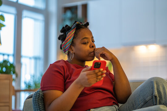 Anxious millenial woman sits on couch at home with phone feels nervous while reading online news or personal message. Upset black female looks uneasy while scrolling smartphone, online stress, tension - Powered by Adobe