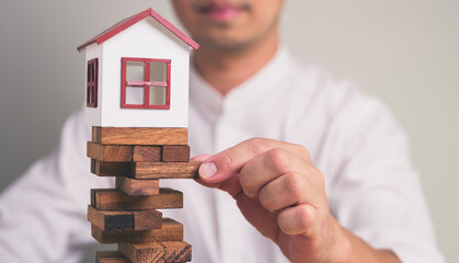 Businessman's hand removing a wooden block from a stack with small model house on top, uncertainty,...