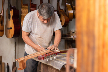 A Latino craftsman stands while measuring a guitar with a tape measure to make another acoustic guitar in a workshop in Neiva, Huila, Colombia. Concep of carpentry