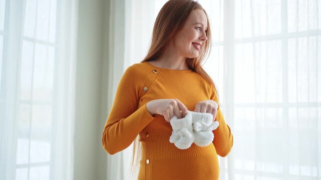 Pregnancy motherhood people expectation future. Pregnant woman with big belly holding newborn baby booties smiling at home. Young mom enjoying pregnancy. Maternity tenderness parenthood new life