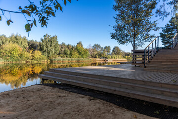 Scenic Lakeside View with Wooden Deck, Capturing the Beauty of Nature Reflected in the Calm Waters Under a Clear Blue Sky Surrounded by Lush Trees and Vibrant Autumn Colors