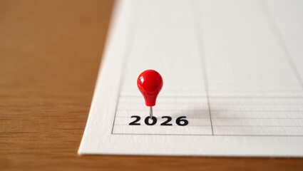 Close-up of a red push pin pointing to the numbers 2026 on white paper, symbolizing future planning, goals, and important dates. The background is blurred to emphasize focus