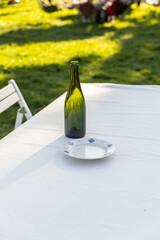 A Serene Outdoor Dining Setup Featuring a Green Bottle on a White Tablecloth Beside a Decorative Plate, Ideal for Summer Gatherings and Relaxing Meals in Nature's Embrace