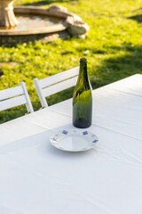 A Charming Outdoor Dining Setup Featuring a Green Bottle and Decorative Plate on a White Table Amidst Lush Greenery Near a Decorative Fountain