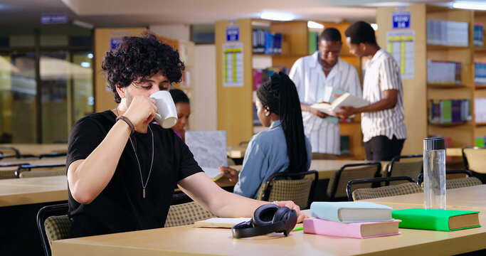 Indian young adult gen z busy male man sitting on chair read book indoor library campus place. Teen pupil boy hold cup mug drink coffee do self exam study notes new skill work break day time - Powered by Adobe