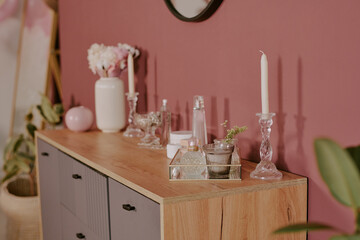 Wooden sideboard holding glassware, candles, vases with flowers and decorative items arranged for bachelorette party celebration, pink wall and plants in background