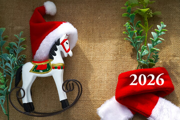  Festive image of a Santa hat placed on a wooden rocking horse, accompanied by vibrant greenery and the year 2026. Perfect for holiday and New Year-themed designs and decorations