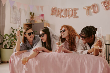 Group of four young adult women wearing sunglasses, laughing and toasting with champagne glasses while celebrating bachelorette party indoors with festive decorations