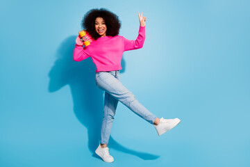 Cheerful girl in casual pink outfit holding colorful skateboard and showing peace gesture against vibrant blue background