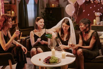 Group of young adult women celebrating bachelorette party, sitting together on sofa, bride wearing veil pouring champagne into glasses, smiling and laughing