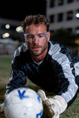 An attentive goalkeeper lies on the grass, ready to save the soccer ball, with floodlights shining brightly, emphasizing the thrilling atmosphere of the night game.