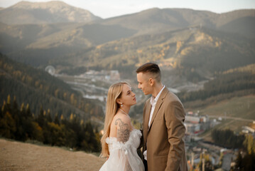Naklejka premium A beautiful, happy young couple in love on a wedding walk and photo shoot high in the mountains. The groom in a jacket and suit and the bride in a white dress at a resort. Kissing and hugging