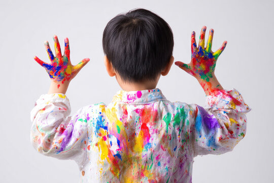 Young Asian child with short black hair, wearing white shirt covered in colorful paint, raises hands showing palms painted in vibrant colors, expressing creativity and joy