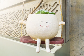 Charming planter with smiling face is hanging on swing, holding vibrant green plants. Playful decor, modern urban environment. Sunlight warm glow, delightful scene of indoor gardening.
