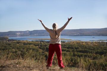 senior woman with gray hair  stands on the mountain with his arms open to the sides on a sunny...